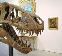 A T. rex skull on a stand with an ornate mirror and a painting on the wall behind it.