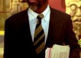 Photograph of Leonardo Patterson, a black man who is aged about 50 wearing a suit and tie holding papers. There are ancient Maya sculptures behind him.
